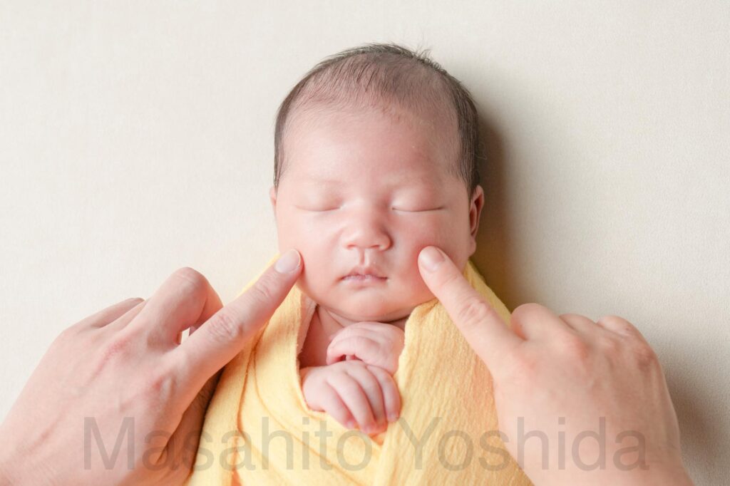 下伊那郡松川町のニューボーンフォト出張撮影|黄色いおくるみ、白い背景でパパとママにほっぺたをツンツンされている赤ちゃんの写真